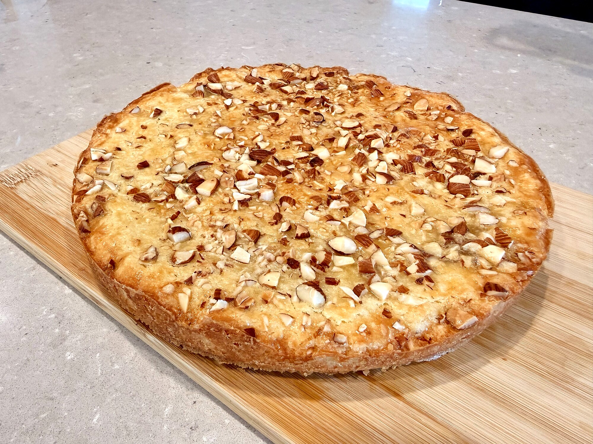 A whole boterkoek on a wooden cutting board sitting on a marble counter, deep golden surface scattered with coarsely chopped whole almonds, edges caramelised to a darker amber, the cake about an inch deep with a soft rumpled top