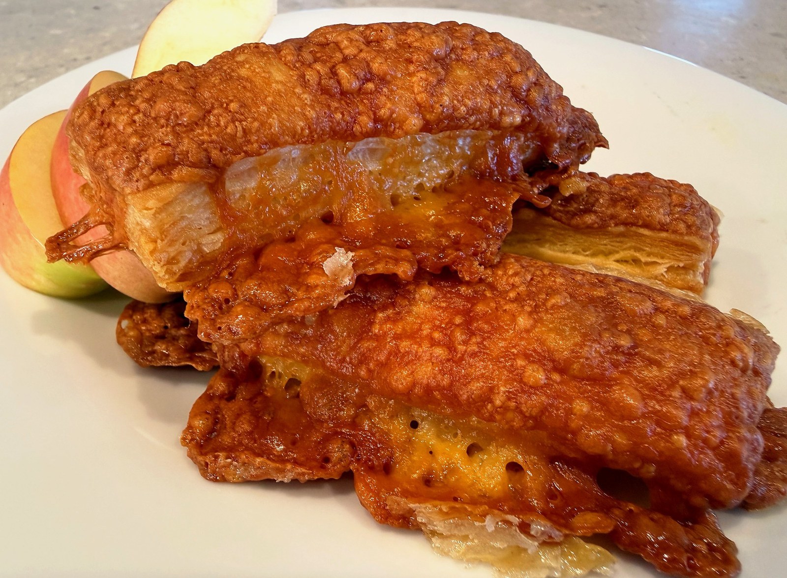 Deep-gold cheese twists on a white plate, layers of pastry visible at the cut ends, melted parmesan caramelised along the seams, sliced apple alongside