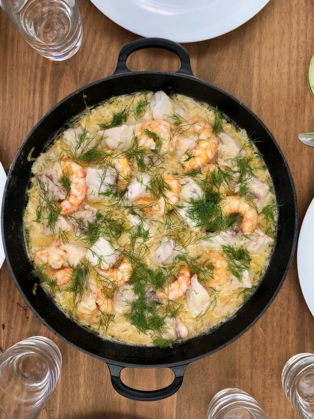 A wide black cast iron pan top-down on a wooden table, full of cod chunks and pink prawns sitting in a pale glossy sauce, scattered all over with feathery green fennel fronds, glasses and plates ringing the edge of the frame