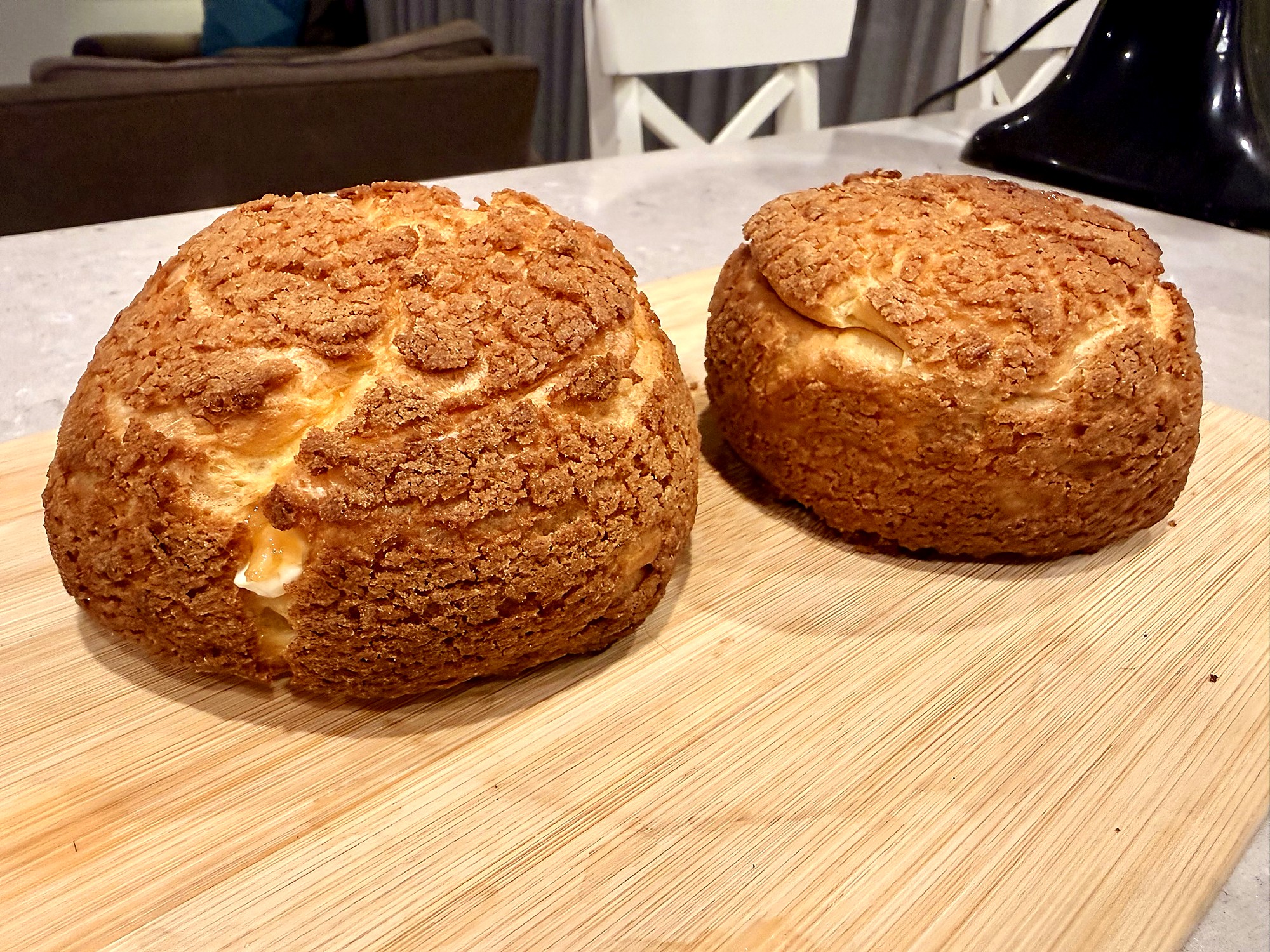 Two large choux buns on a wooden cutting board, each capped with a craquelin crust crazed into a crackled golden-brown lid, the choux pastry beneath showing soft cream filling pushing through the cracks