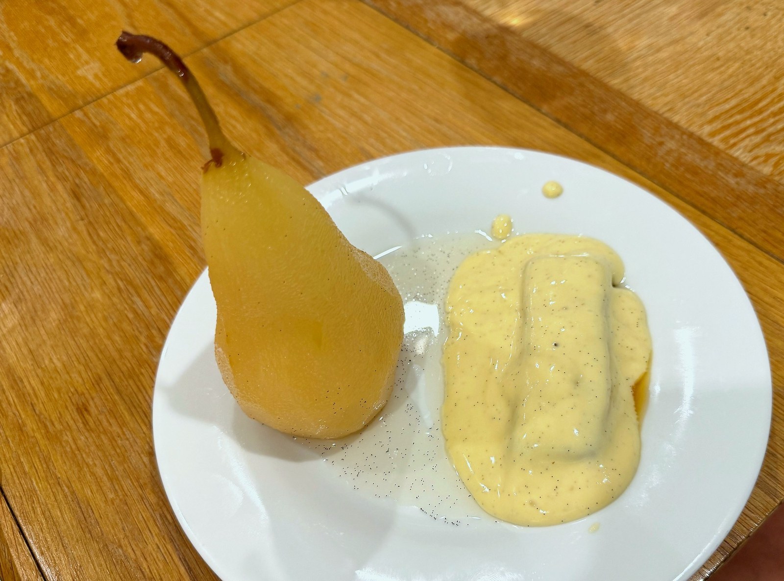 A whole poached Beurre Bosc on a white plate, pale gold and vanilla-flecked, served alongside a pool of crème pâtissière