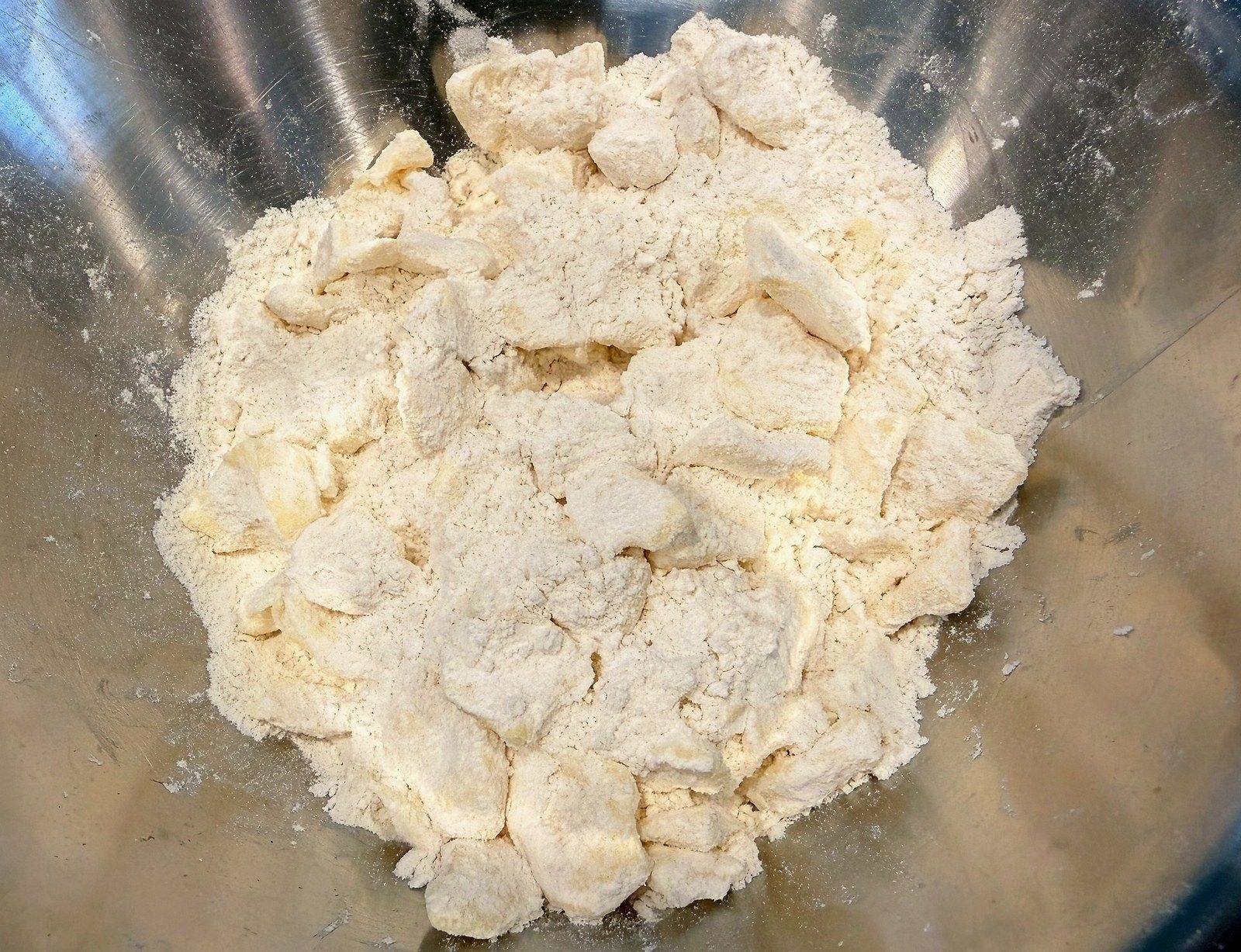 Butter cubes tossed through flour in a metal mixing bowl, each cube whitened with a coating of flour before being pressed flat between finger and thumb